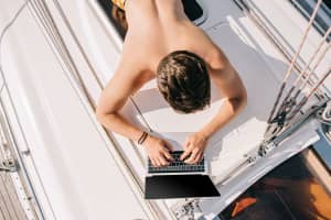 A young boater working on a laptop on a sailboat deck, calculating daily boat electrical loads and amp-hour requirements